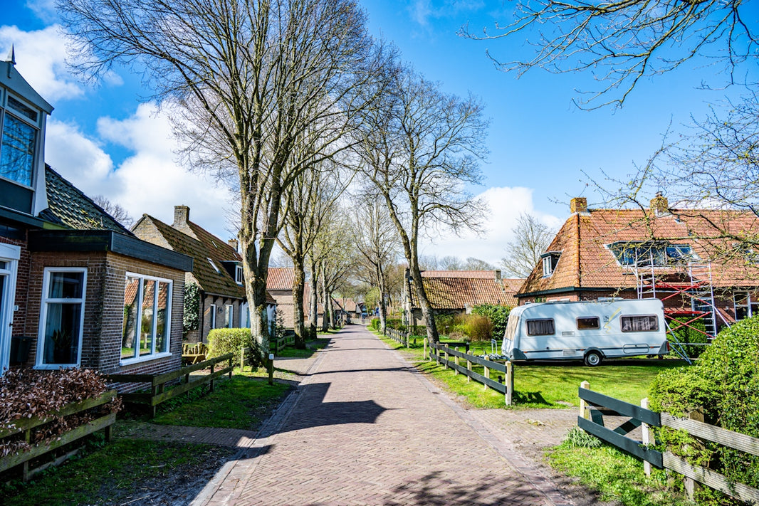 Sunny village street with houses, trees, and a parked caravan.