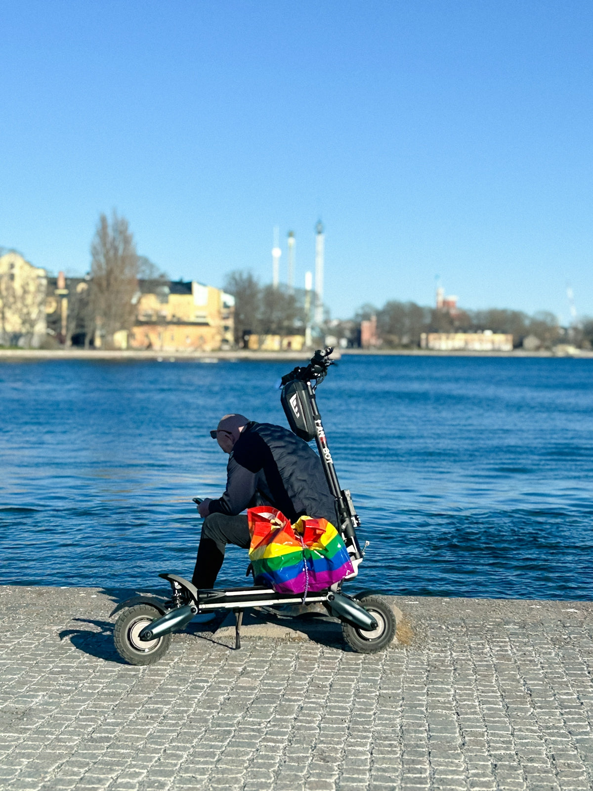 Person on scooter with rainbow bag by calm blue water
